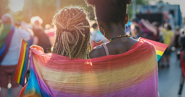two individuals with a rainbow flag draped across their shoulders for Pride Month
