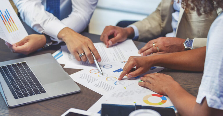 closeup image of table with paperwork, laptop, and colleagues working